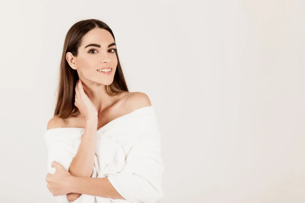 Woman with long brown hair wearing a white off-the-shoulder top, smiling and touching her neck, against a white backdrop.