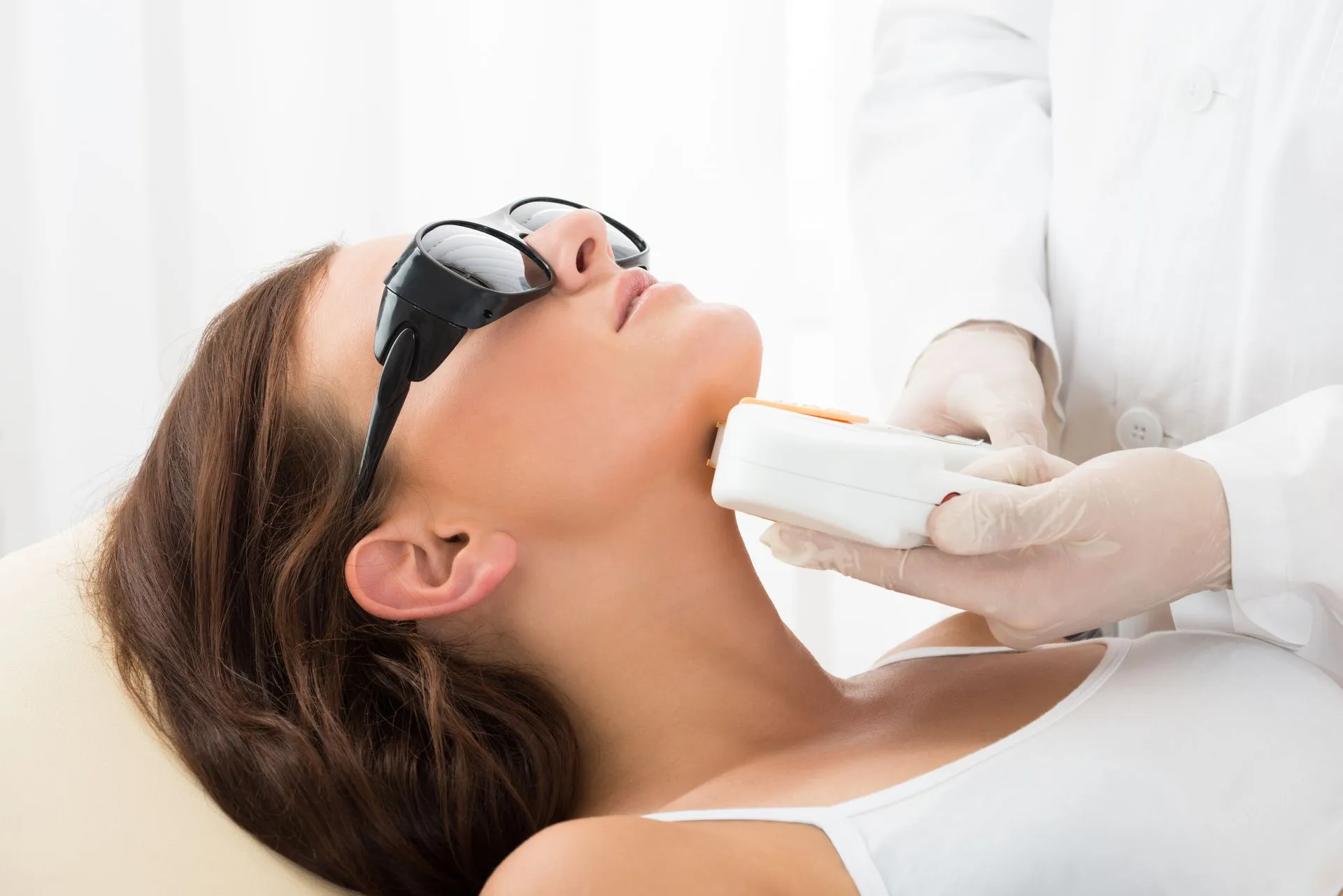 Woman receiving laser hair removal on her neck; wearing protective goggles, doctor in white coat.