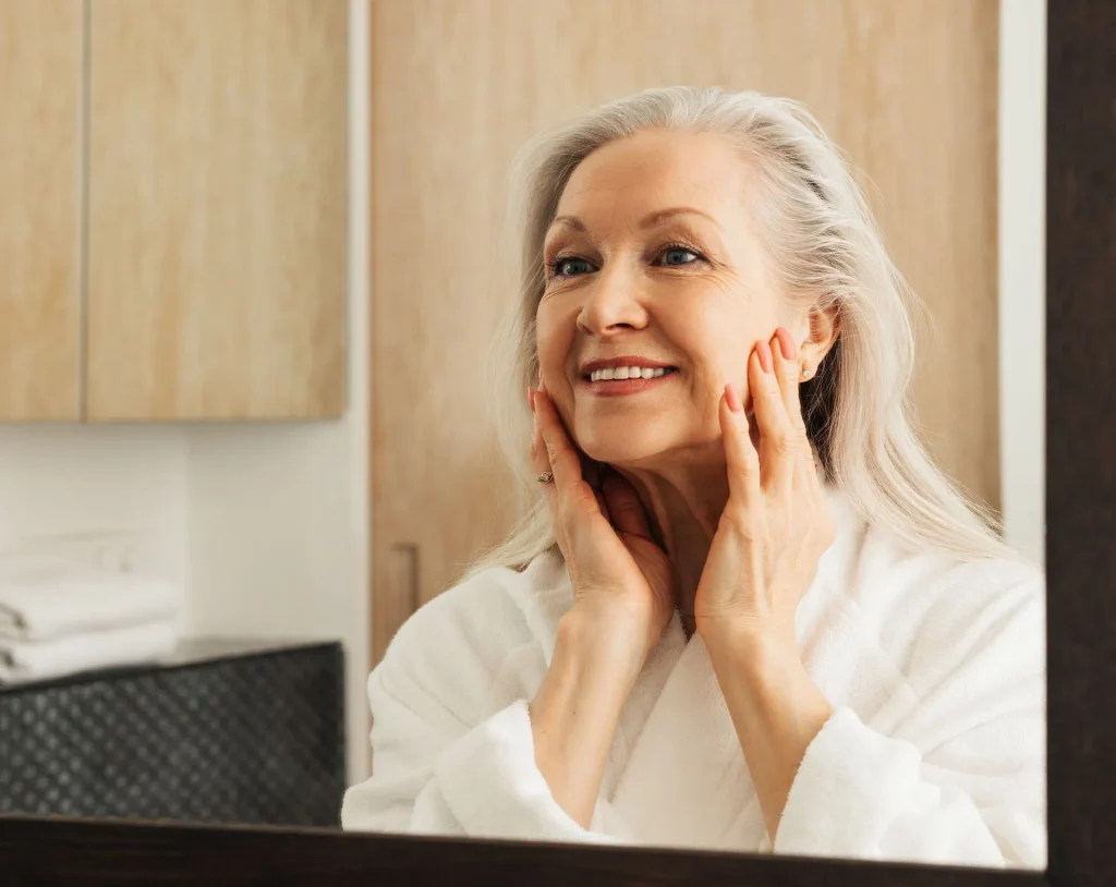 An older woman is looking at her face in a bathroom mirror.